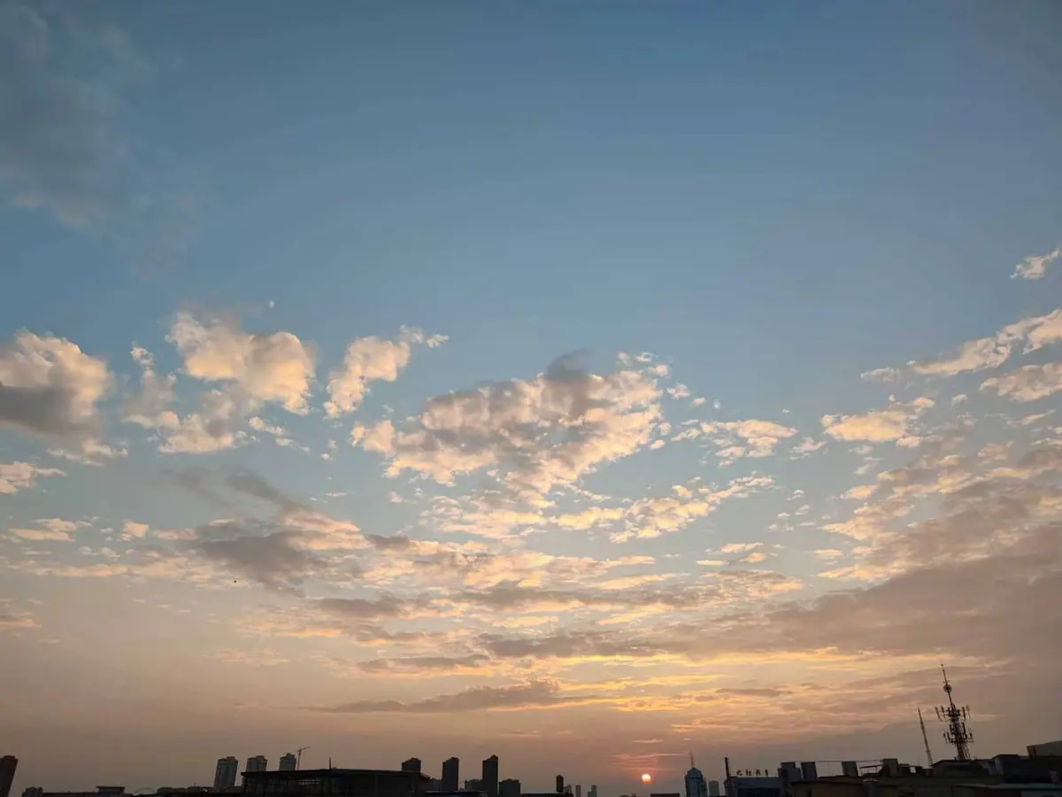 Sunset sky with scattered clouds, the sun near the horizon, and a dark city skyline below.